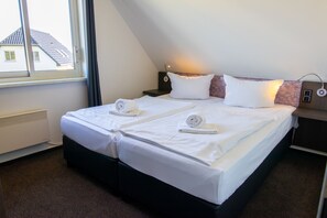 A twin bed with white linens and towels, a bedside table with a lamp, and a window with a view of a house. - Holiday home Type V - Van der Valk Resort Linstow (Dobbin-Linstow)