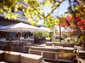 An outdoor seating area with tables and chairs, umbrellas, and a building in the background. - Holiday home Type V - Van der Valk Resort Linstow (Dobbin-Linstow)