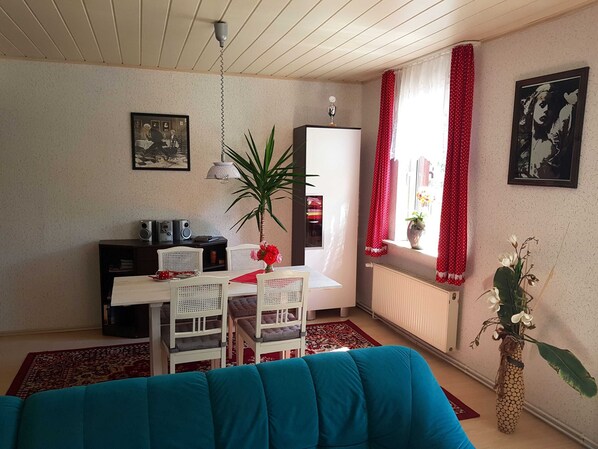 A living room with a sofa, a dining table with chairs, a radiator, a potted plant, and a window with red curtains.