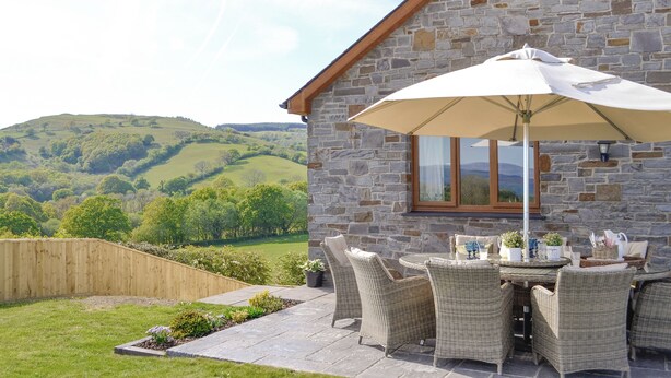 A stone building with a patio area featuring wicker furniture and a table with an umbrella. - Maes Y Bryn Farmhouse (Llansadwrn, near Llandeilo)