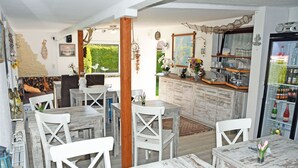 A rustic dining area with wooden tables and white chairs, a kitchen area with a refrigerator stocked with beverages, and a view of greenery outside.