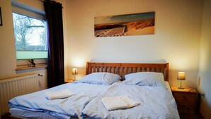 Una cama bien hecha con almohadas y una toalla doblada, una ventana con cortinas, un cuadro colgado en la pared y lámparas de noche.