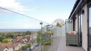 A balcony with a clear view of the sea and houses, a wicker sofa, and a glass railing. - 3 bedroom accommodation in Prestatyn (Prestatyn)