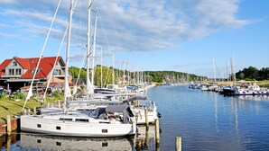 En lysthavn med flere sejlbåde fortøjet ved moler, et hus med rødt tag, og stille vand der spejler både og himmel.