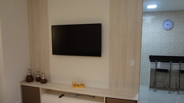 Una sala de estar moderna con un televisor de pantalla plana montado en la pared, un estante con objetos decorativos y una zona de cocina con un reloj en la pared.