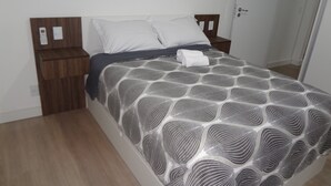 Una cama bien hecha con una colcha estampada, una mesita de noche de madera con un teléfono e interruptor de luz, y una puerta a la derecha.