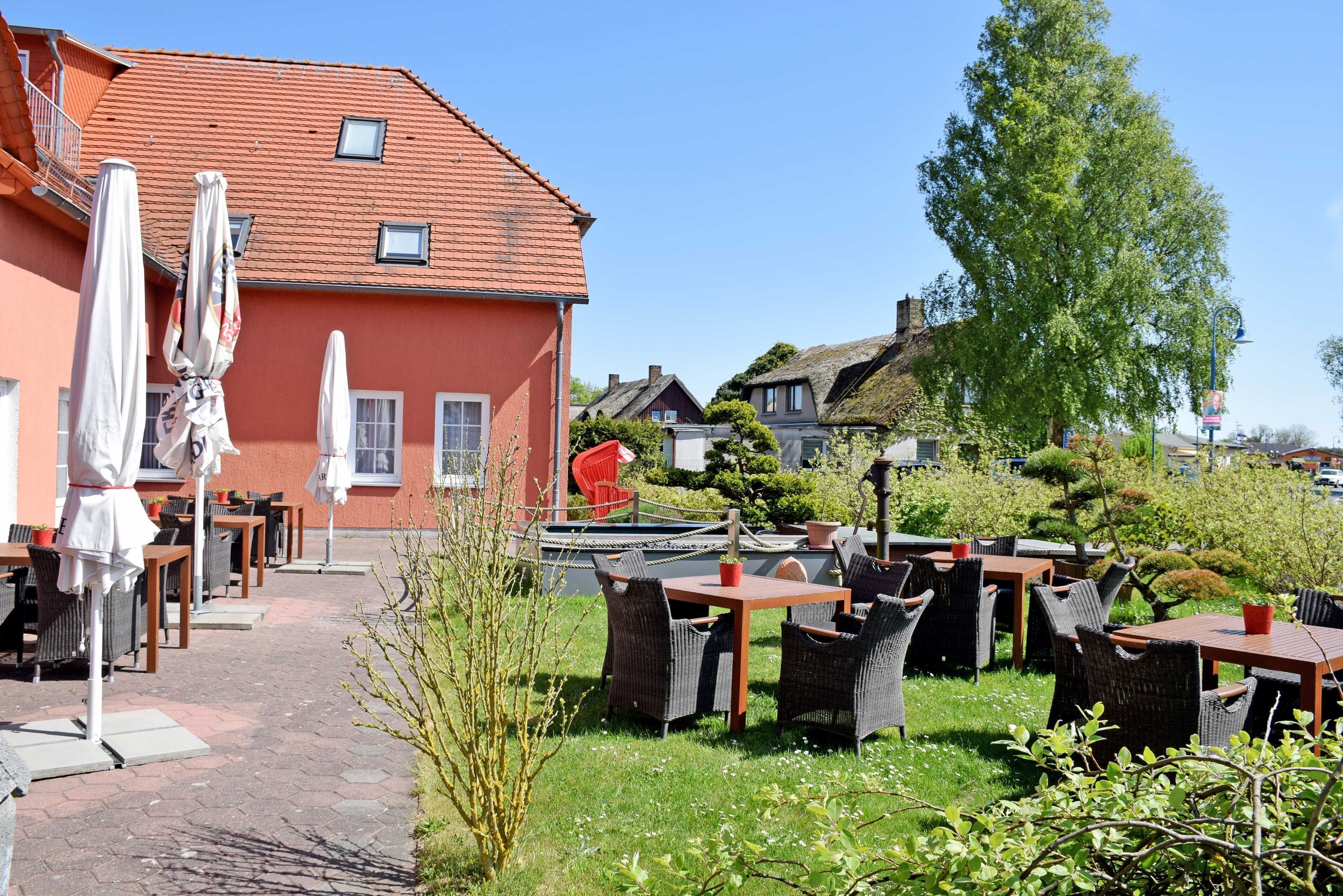 An outdoor cafe with tables and chairs, a red building in the background, and a tree.