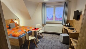 Una habitación de hotel compacta con una cama, una mesita con una silla, un escritorio con una silla, un radiador, una ventana con cortinas y un televisor montado en la pared.