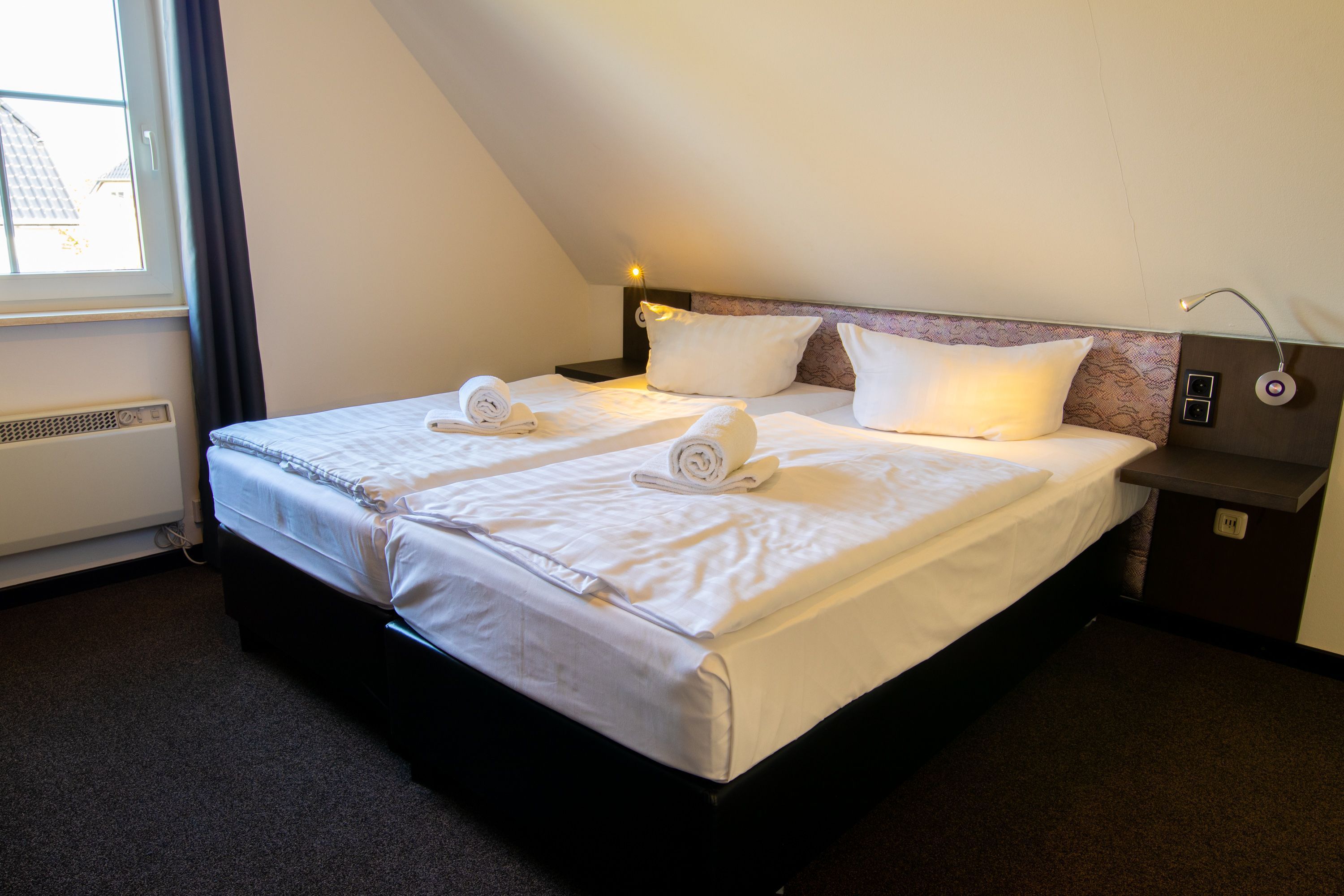 Una habitación de hotel con dos camas, una ventana, un calentador y una mesita de noche con lámpara.