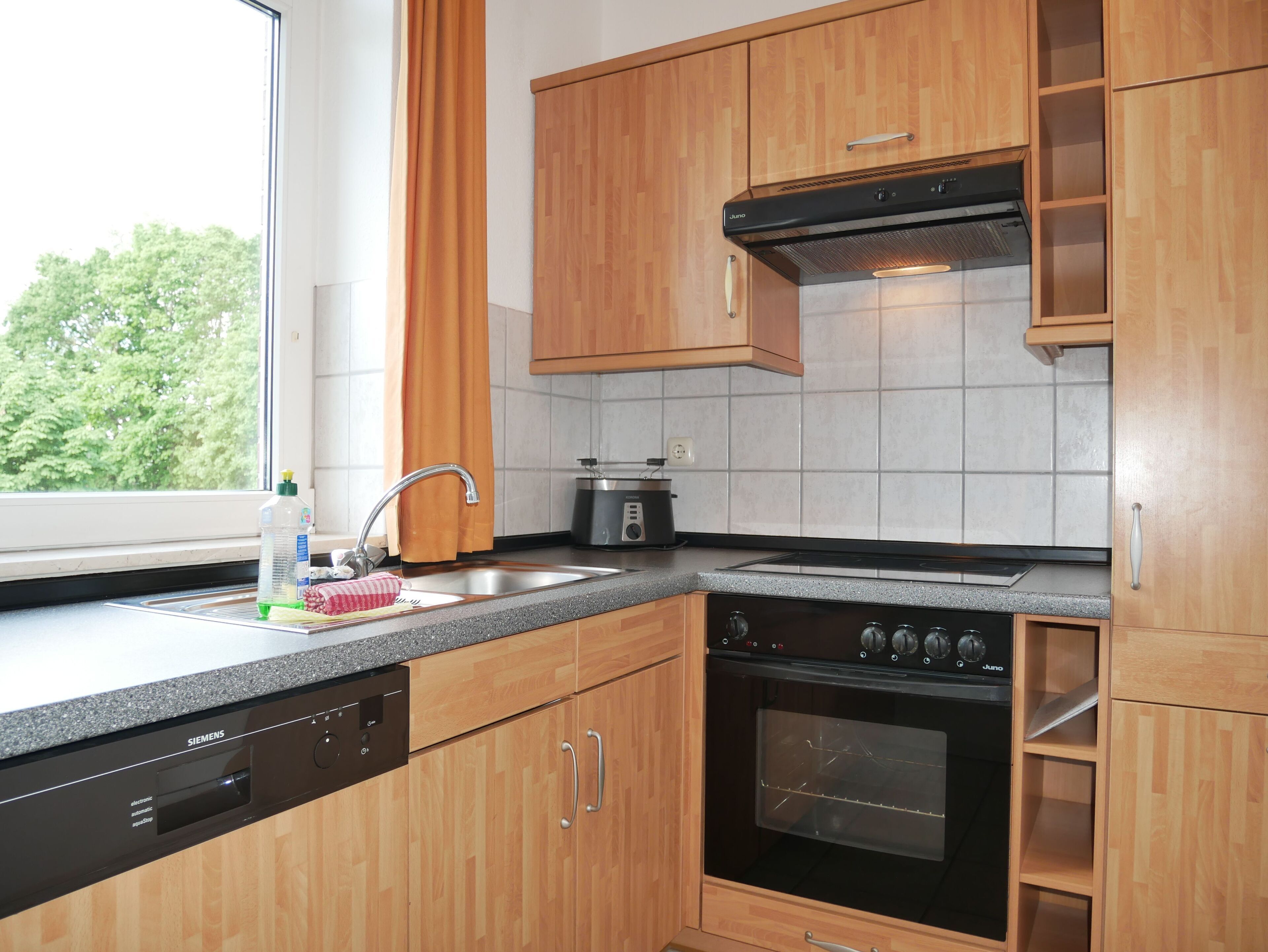 A modern kitchen with wooden cabinets, a built-in oven, a dishwasher, and a window with a view of trees.