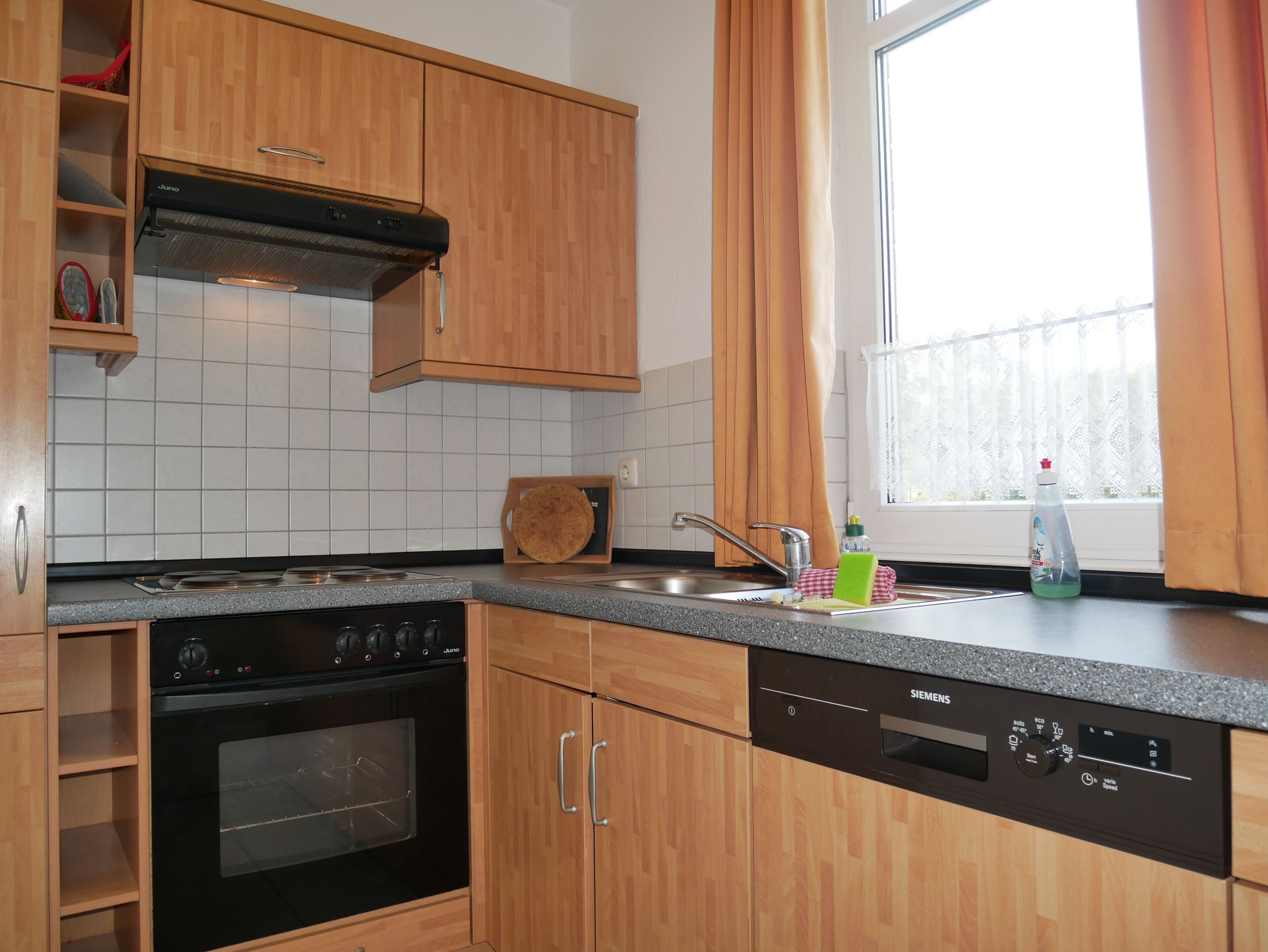 Een moderne keuken met houten kastjes, een RVS spoelbak, een vaatwasser en een raam met gordijnen.