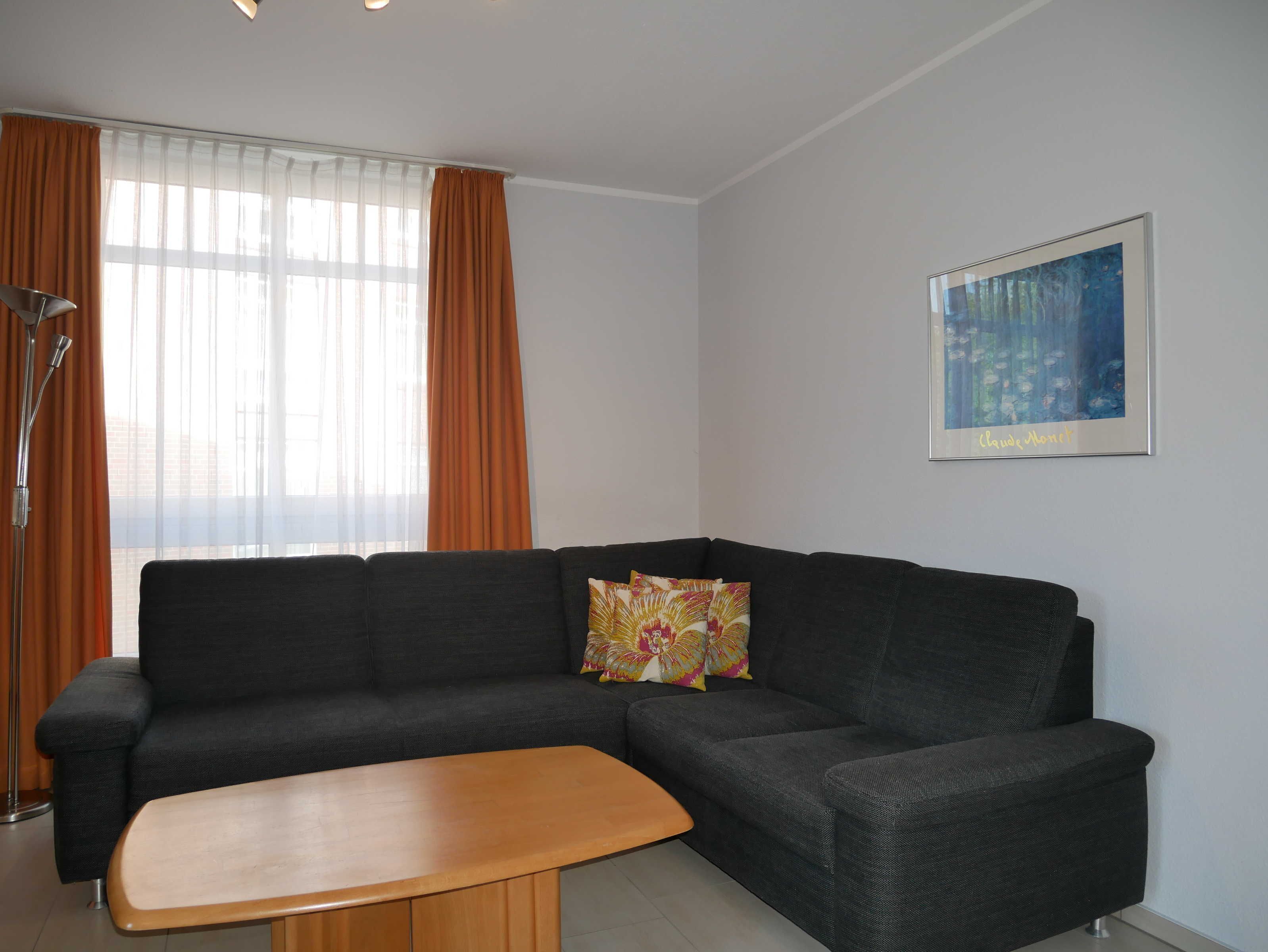 A modern living room with a dark grey sectional sofa, a wooden coffee table, and a large window with orange curtains. A framed abstract painting is on the wall.