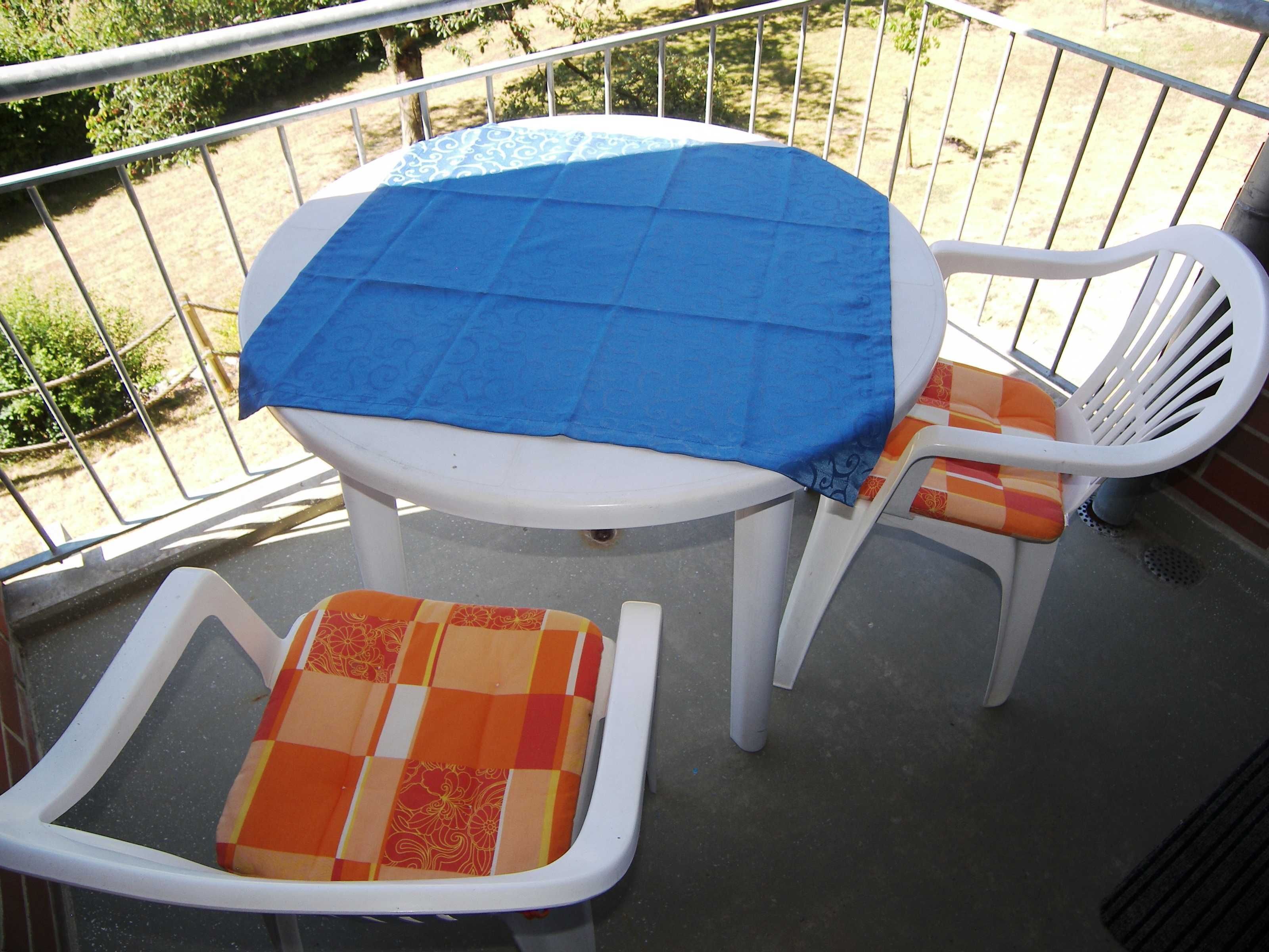 A small outdoor balcony with a round table covered by a blue tablecloth, two white chairs with orange cushions, and a metal railing.