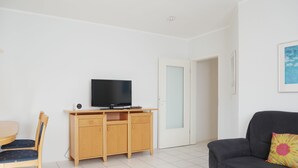Een moderne woonkamer met een flatscreen televisie op een houten kast, een eethoek met tafel en stoelen, een bank met een kleurrijk kussen, en een deur die leidt naar een andere kamer.