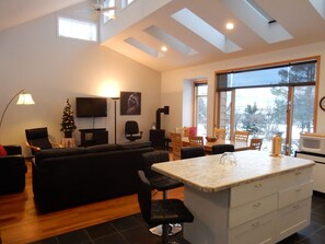 A modern living room with a black sofa, a coffee table, and a TV. There is a dining area with a table and chairs, and a kitchen area with a microwave.
