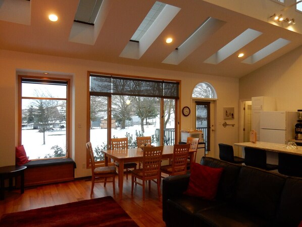 A spacious living area with a wooden dining table and chairs, a black leather sofa, and a large window offering a view of a snowy landscape.