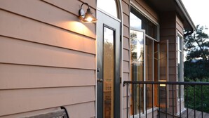 Una terraza de madera con un banco, baranda metálica y un edificio con ventanas amplias y una puerta.