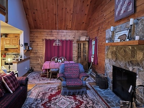 A cozy log cabin interior with a dining area, a fireplace, and a kitchen visible through an open door. - Mt. Snow/Stratton Log Cabin in the Woods (West Wardsboro)
