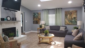 A living room with a fireplace, a flat-screen TV, a sofa, a coffee table, and two lamps. - Sharnu's Palace (DeSoto)