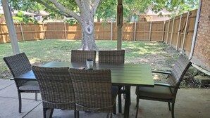 An outdoor patio with a table and chairs, a tree, and a wooden fence in the background. - Sharnu's Palace (DeSoto)
