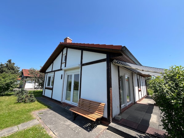 Une maison à deux étages, avec une façade blanche et des éléments décoratifs sombres, un banc en bois à l’extérieur et un ciel bleu dégagé.