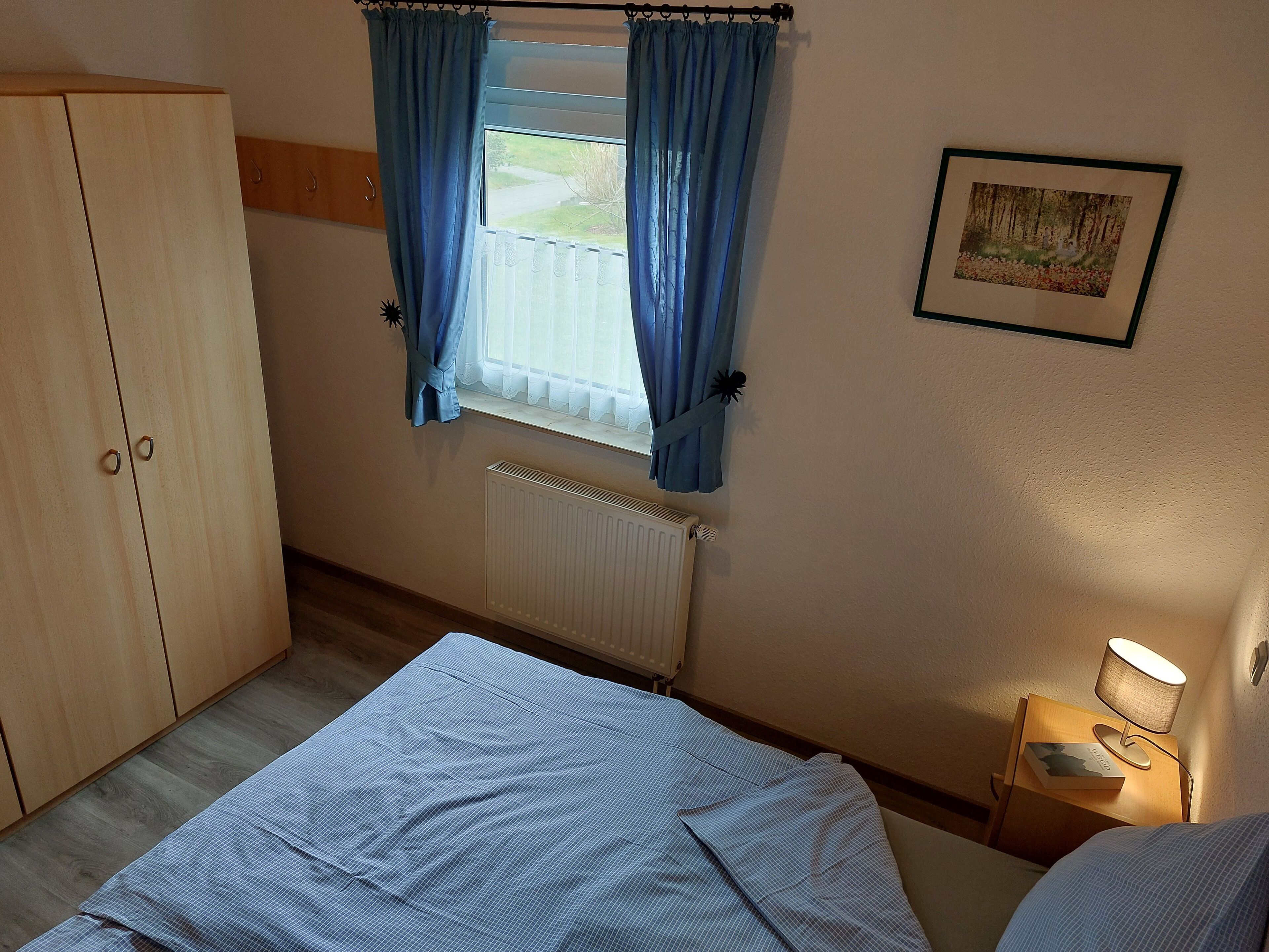 Une chambre à coucher comprenant un lit, une armoire, une fenêtre avec des rideaux, un radiateur, une table de chevet avec une lampe et un tableau encadré accroché au mur.
