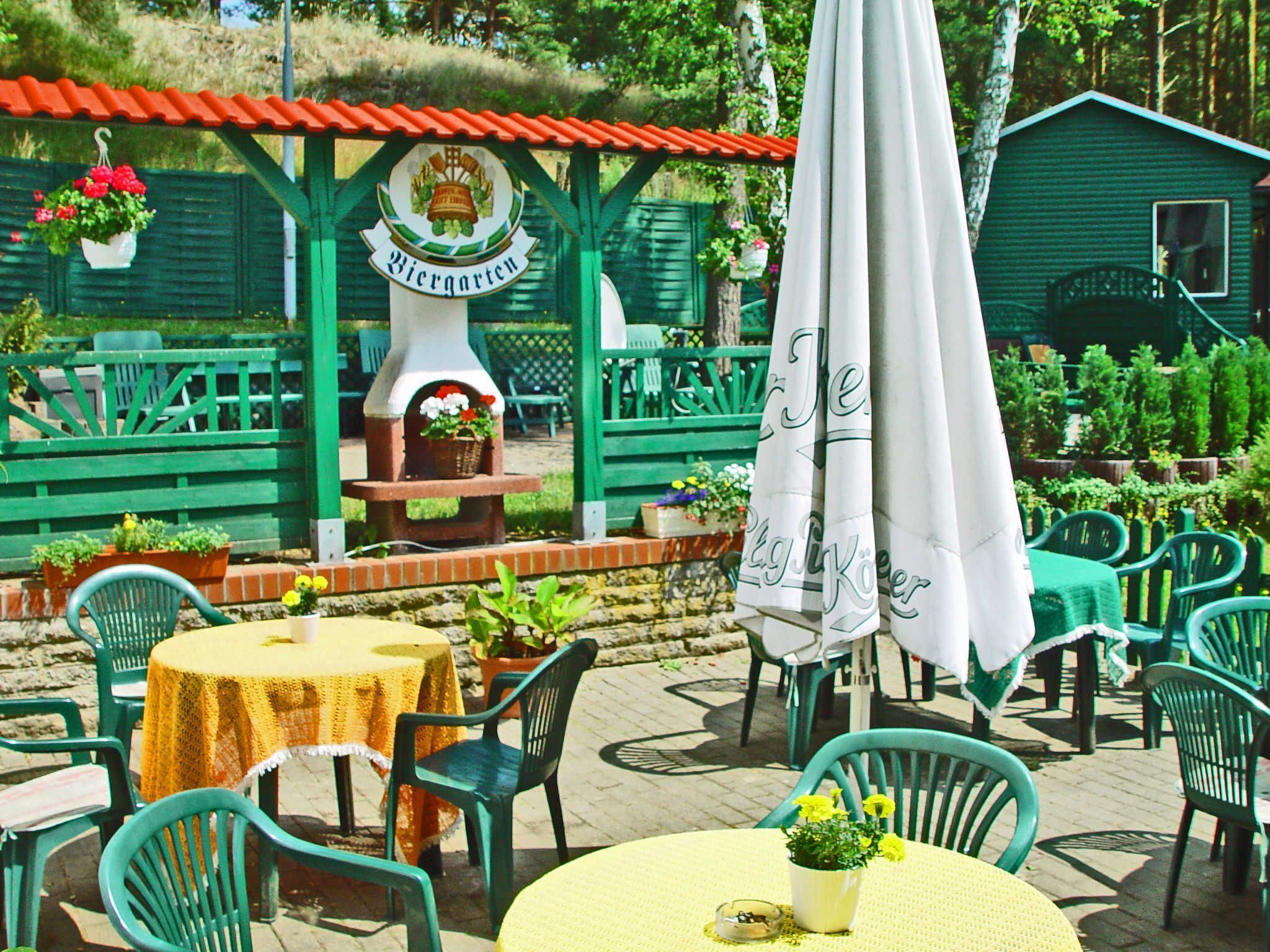 Un espace extérieur aménagé avec des chaises et des tables vertes, un panneau indiquant un jardin à bière et un four en briques.