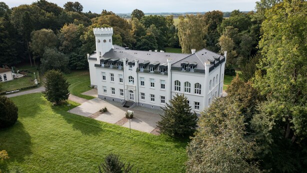 Een groot, wit, kasteelachtig gebouw met veel ramen en een toren, omringd door weelderige begroeiing en een verzorgd gazon.