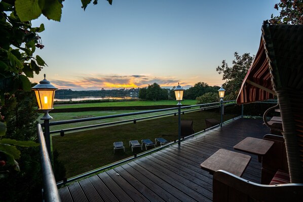 A deck with outdoor seating and two lit lanterns, overlooking a grassy area and a sunset view. - Double room - Hotel Am Gothensee (Heringsdorf)