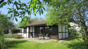 A small, single-story building with a tiled roof, a patio with chairs, and a lawn.