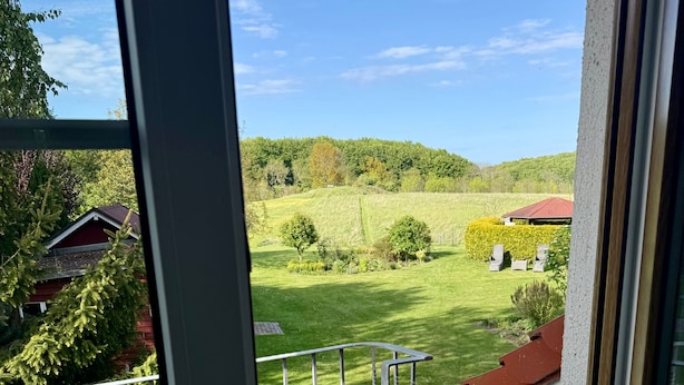 Ein Balkon mit Blick auf eine grüne Landschaft, darunter Bäume und ein Gebäude mit rotem Dach.
