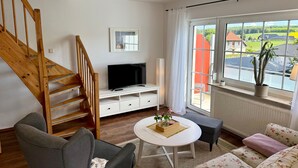 Ein Wohnzimmer mit einer Holztreppe, einem Flachbildfernseher, einem Sofa, einem Couchtisch und einem Fenster mit Blick auf Häuser und Grünflächen.