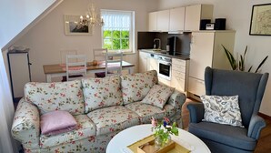 Ein modernes Wohnzimmer mit einem blumigen Sofa, einem grauen Sessel, einem runden Couchtisch mit einer Vase, einem Essbereich mit Tisch und Stühlen sowie einer Küche mit Schränken und Geräten.