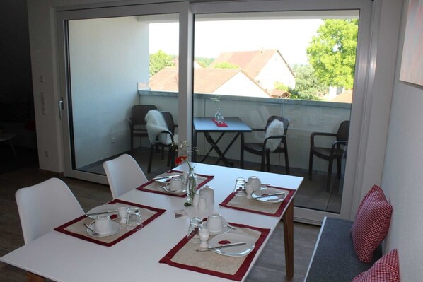 A dining area with a table set for four, chairs, a small table with a vase, and a view of the outside.