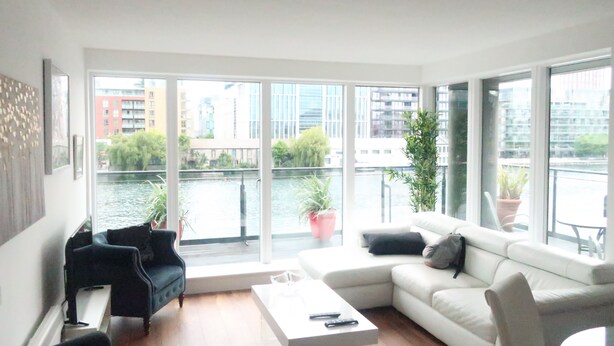 A modern living room with a white sofa, a black armchair, and a coffee table. Large windows offer a view of a river and buildings. - Grand Canal Dock Waterfront Luxury “Special Offer” (Dublin)
