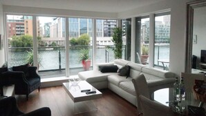A modern living room with a large window overlooking a body of water and buildings. There is a white sofa, a glass coffee table, and a black armchair. - Grand Canal Dock Waterfront Luxury “Special Offer” (Dublin)