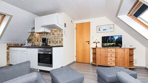 A compact living space with a kitchenette, grey upholstered seating, a wooden cabinet, and a television. - Apartment 8 - Haus Störtebeker (Neuharlingersiel)