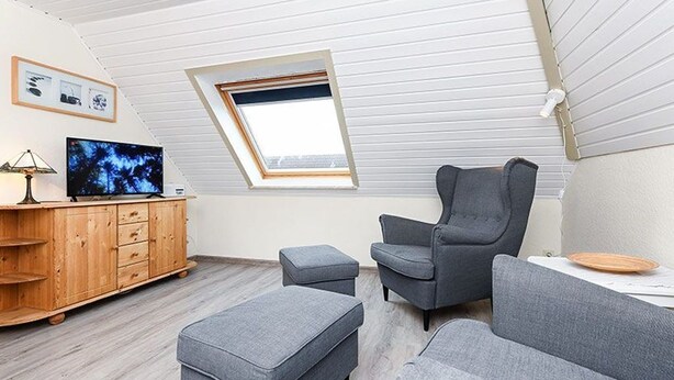 A modern loft bedroom with a sloped ceiling, a wooden wardrobe, a grey sofa, a grey armchair, and a television on a wooden stand. - Apartment 8 - Haus Störtebeker (Neuharlingersiel)