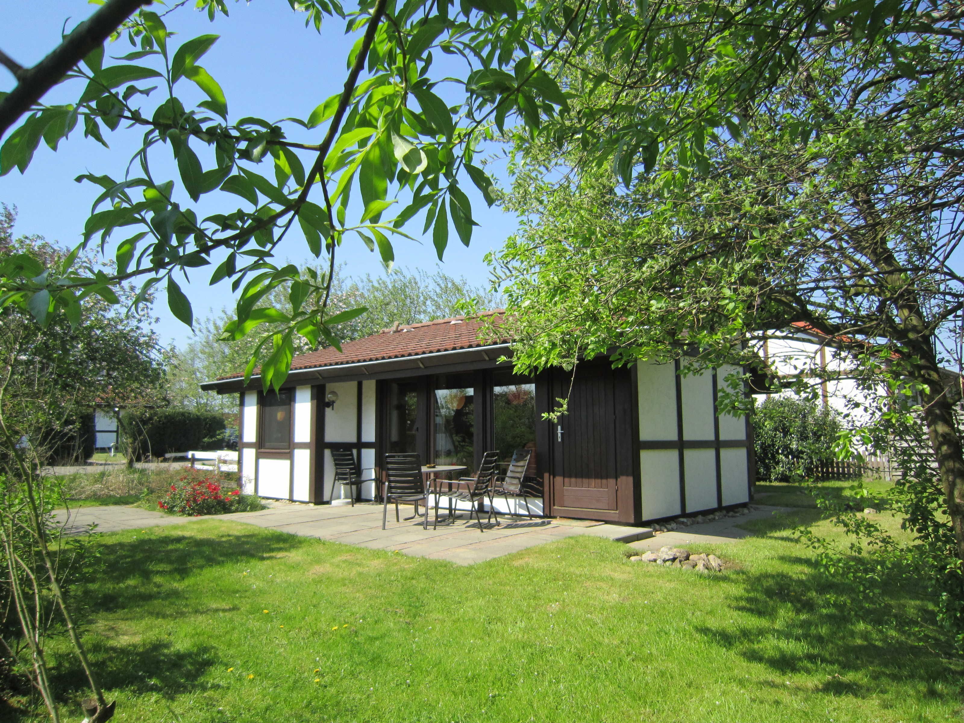 Ein kleines, eingeschossiges Gebäude mit Ziegeldach, einer Terrasse mit Sitzgelegenheiten und einem Rasen.