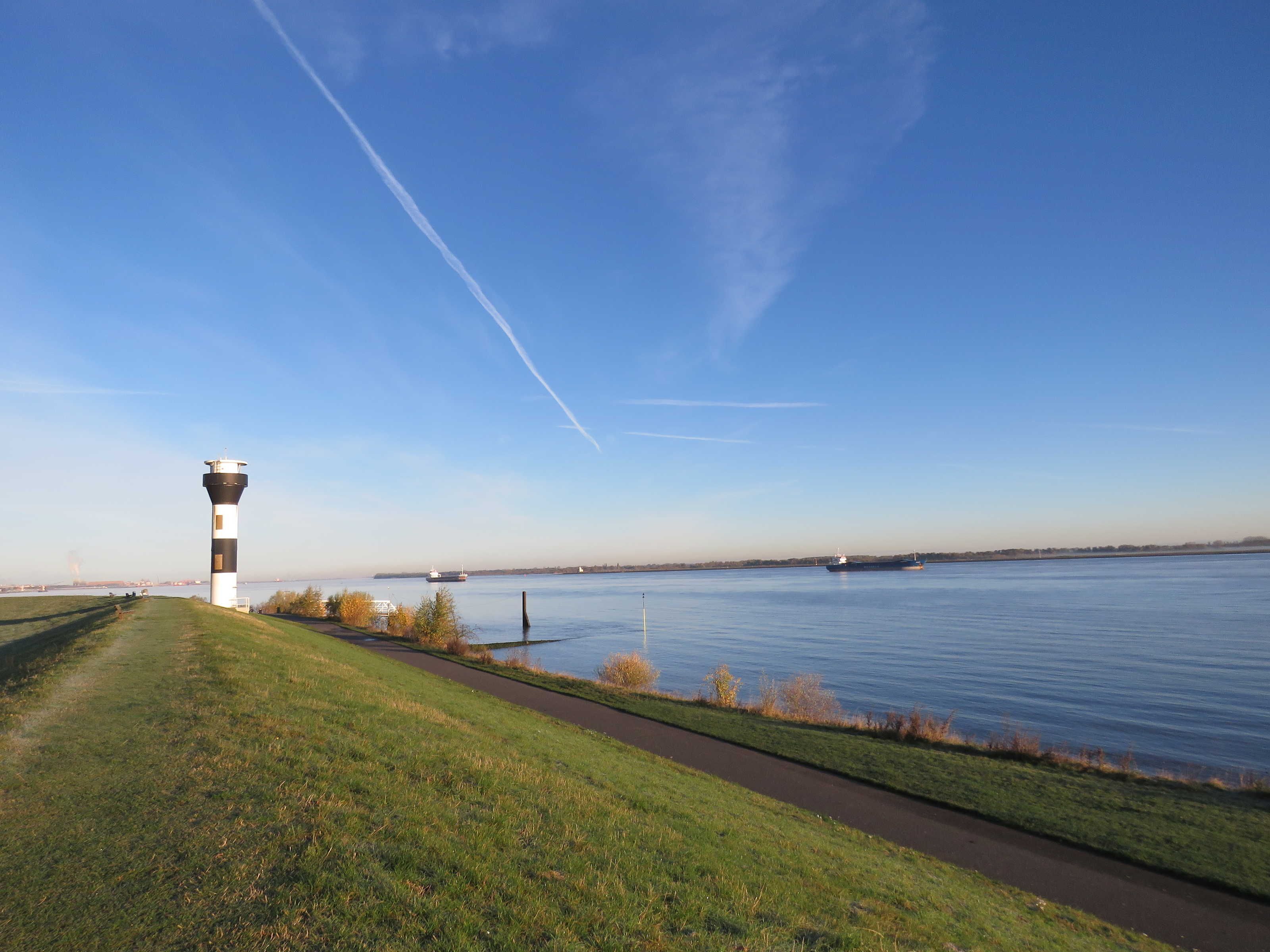 Un phare se dresse sur une colline herbeuse, surplombant une étendue d’eau calme sous un ciel dégagé.