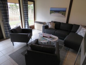 Uma sala de estar com sofá, poltronas e mesa de centro. Há um quadro na parede e uma janela com cortinas.
