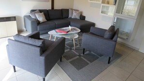A modern living room with a grey sofa, two armchairs, a coffee table, and a rug. There is a painting of a beach on the wall, a small plant on a shelf, and a cabinet with glass doors.