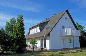 Uma casa de dois andares com varanda, telhado inclinado e um jardim com plantas em vasos.