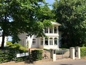 A white building with a balcony, surrounded by trees and a metal fence.