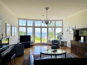 Ein geräumiges Wohnzimmer mit großem Fernseher, bequemen Sitzgelegenheiten und Meerblick durch große Fenster.