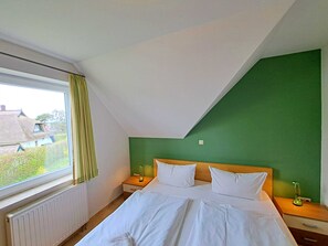 A bedroom with a large bed, two bedside tables, a window with curtains, and a view of a house outside.