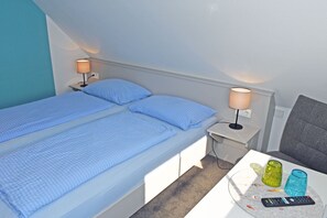 Een tweepersoonsbed met een blauwe spreideken en kussens, een nachtkastje met een lamp, een klein tafeltje met een afstandsbediening, en een stoel.