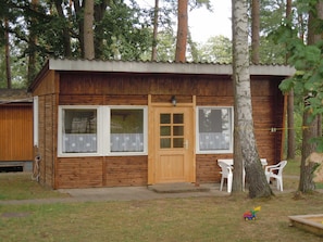 A wooden cabin with a front door, two windows, and a small porch with plastic chairs. - Bungalow 3 - Ferienanlage Am Sidowsee (Fürstenberg/Havel)