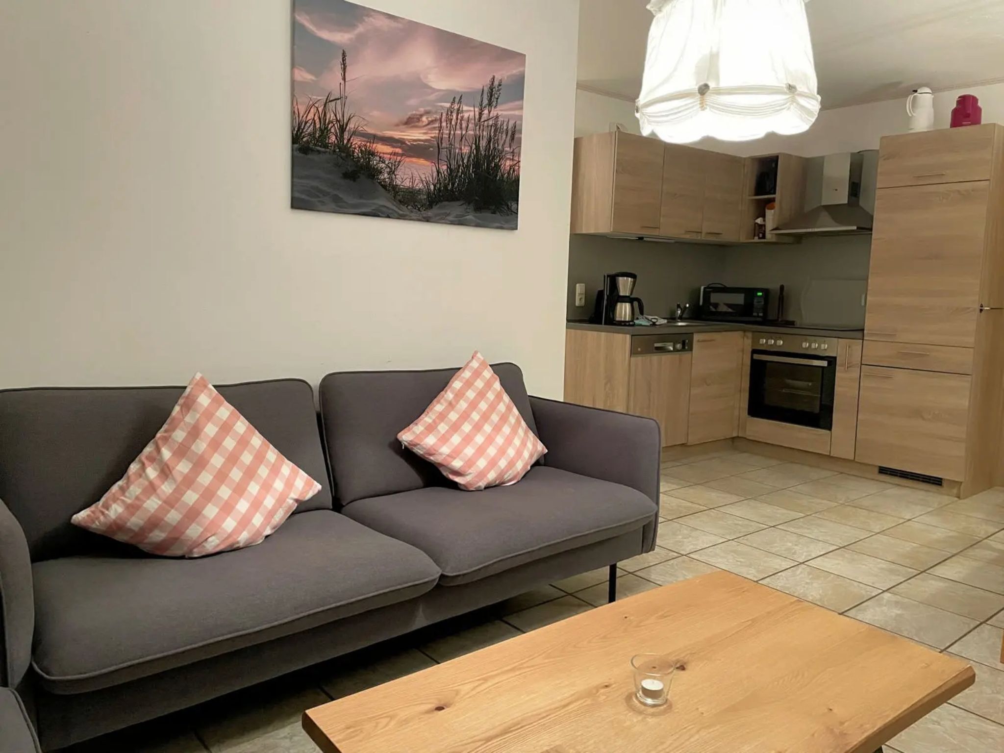 Ein modernes Wohnzimmer mit grauem Sofa und karierten Kissen, einem Holztisch und einer Küchenzeile mit Holzschränken und Geräten.
