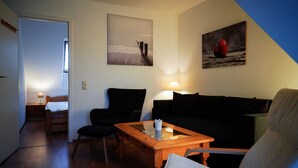 Un salon chaleureux avec une table basse en bois, un canapé noir et un fauteuil. Un tableau représentant un cœur est accroché au mur, ainsi qu’un autre tableau représentant une jetée.